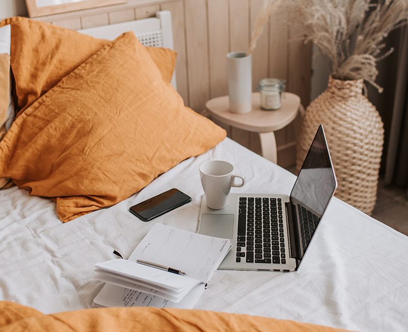 Room decoration of a laptop, phone, notebook and mug are placed on white bedsheets against orange pillows in a los angeles home